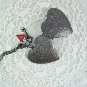 May include: A silver locket in the shape of a heart with an intricate design. The locket is open and has a red heart charm attached to it with a silver chain.