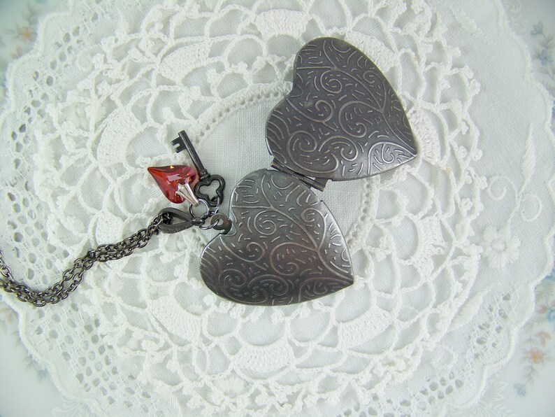 May include: A silver locket in the shape of a heart with an intricate design. The locket is open and has a red heart charm attached to it with a silver chain.