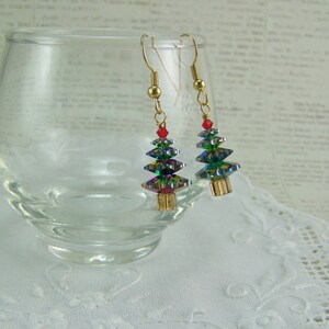 May include: A pair of Christmas tree earrings. Each earring features a gold-colored hook, a red bead, and a tree-shaped design made of iridescent, faceted crystals. The earrings are displayed near a clear glass and a white lace doily.