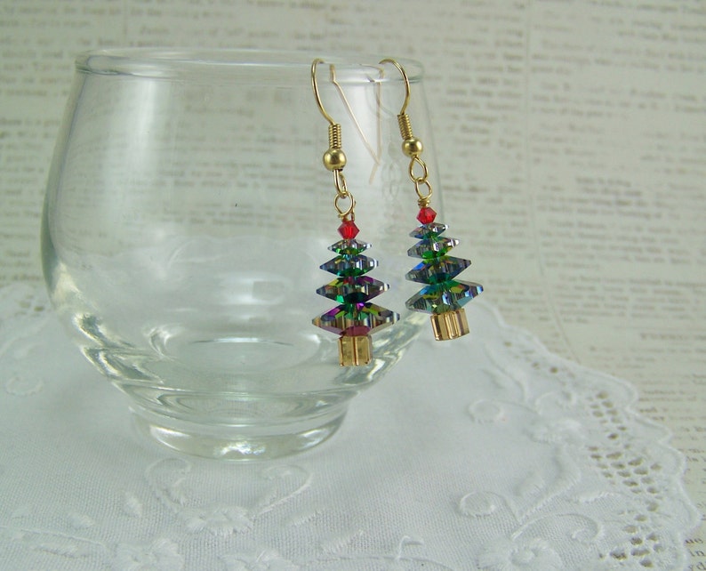 May include: A pair of Christmas tree earrings. Each earring features a gold-colored hook, a red bead, and a tree-shaped design made of iridescent, faceted crystals. The earrings are displayed near a clear glass and a white lace doily.