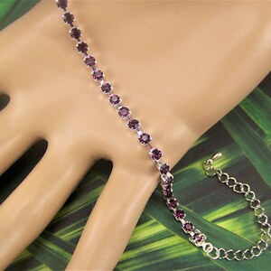 May include: A silver-toned bracelet featuring a line of round, deep purple gemstones. The bracelet is shown on a light-skinned hand against a backdrop of green and yellow foliage. The bracelet has an adjustable chain.