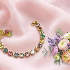 May include: A gold-tone bracelet featuring a series of round, multi-colored faceted crystals in shades of pink, yellow, blue, and green. The bracelet includes a gold-tone lobster clasp and a small gold-tone leaf charm attached to one end.