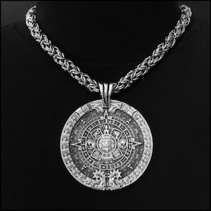 May include: A silver chain necklace with a large, round pendant featuring a detailed design of the Aztec calendar. The pendant is intricately carved with symbols and figures.