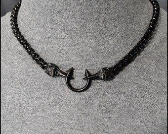 May include: A black chain necklace with a black metal ring and two pointed ends. The chain is braided and has a textured surface.