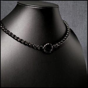 May include: A black metal chain necklace featuring a series of interlocking links and a central circular ring component. The necklace has a polished, dark finish and is displayed on a smooth, black neck form to showcase its structure and design.