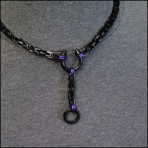 May include: A black chain necklace with a black metal pendant featuring a ring and two spikes. The chain has purple accents.