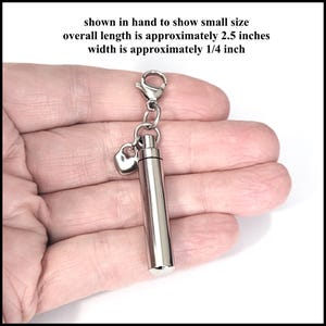 May include: A silver-colored, cylindrical pendant with a small heart charm attached. The overall length is approximately 2.5 inches, and the width is approximately 1/4 inch. The text in the image says "shown in hand to show small size".