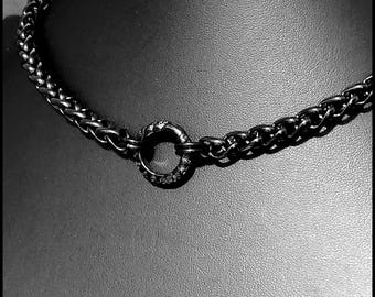 Puede incluir: Un collar de cadena en tono gris metalizado oscuro con eslabones entrelazados de estilo trenzado. El collar está centrado por un componente de anillo circular que muestra un sutil patrón floral o de hojas grabado en su superficie exterior.