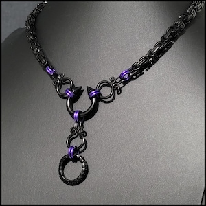May include: A black chain necklace featuring a byzantine link pattern with purple jump ring accents. The centerpiece consists of a black horseshoe-shaped component with two spikes, connected to a dangling black ring via decorative metal scrollwork.