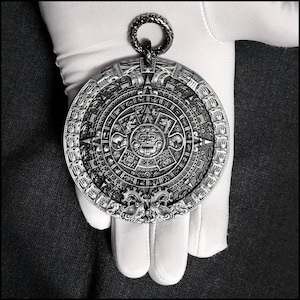 May include: A silver pendant featuring a detailed replica of the Aztec calendar, also known as the Sun Stone. The pendant is circular and has a intricate design with glyphs and symbols.