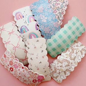 May include: A collection of decorative hair clips with scalloped edges. The clips feature various patterns, including rainbows, floral prints, gingham, bees, bows, and unicorns. The color palette includes pink, blue, green, and white, set against a pink background.