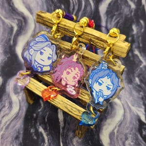 May include: Three acrylic keychains with cartoon character designs. Each keychain has a gold-colored clasp and a small charm. The characters are in blue, pink, and blue, with the keychains hanging on a small wooden bench.