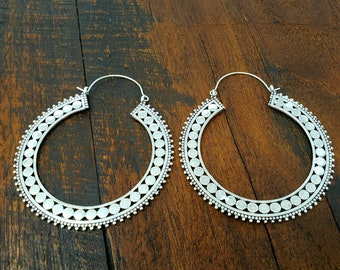 Peut inclure: Une paire de créoles argentées. Les boucles d'oreilles présentent un motif circulaire avec une série de petits cercles espacés régulièrement sur les bords intérieur et extérieur. Sur une surface en bois foncé.
