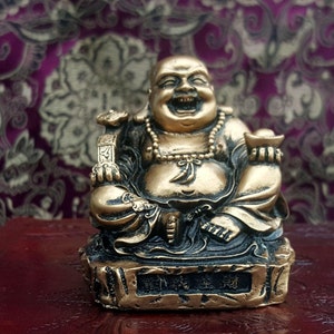 Puede incluir: Una estatua dorada de Buda riendo sentado en una base con caracteres chinos. La estatua está hecha de resina y tiene un diseño detallado.