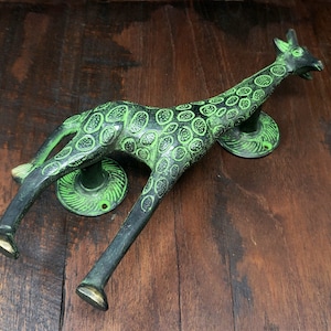 May include: Green and black metal giraffe door knocker with a spotted pattern. The giraffe is lying down with its head turned to the side. The knocker has two round bases.