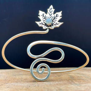 May include: Silver-toned cuff bracelet with a maple leaf design. The leaf, with intricate detailing, is set with a small, round, light-colored stone. The bracelet features a swirling, open design with a spiral at the base.