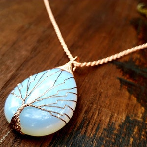 May include: A handcrafted pendant necklace featuring a pear-shaped, opalescent stone with a copper wire tree design. The pendant hangs from a twisted copper chain. The necklace is displayed on a wooden surface.