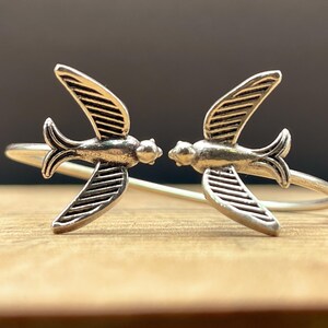 May include: Two silver rings, each with a swallow bird design. The birds face each other, with their wings spread. The rings are set on a wooden surface with a dark background.