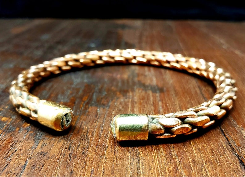 May include: A gold-toned metal bracelet with a woven design. The bracelet is open and has two round, solid metal ends.
