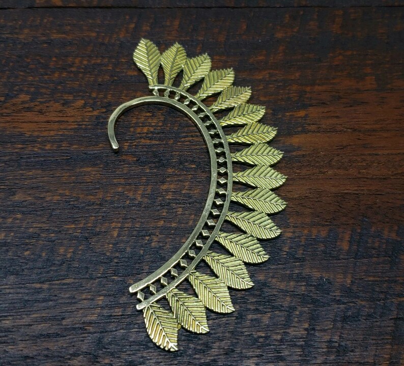 May include: Gold ear cuff with a leaf design. The cuff is curved and has a delicate, intricate pattern.