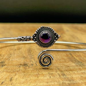 May include: A silver-toned cuff bracelet with a central, round, deep purple gemstone. The gemstone is surrounded by a detailed, beaded setting. The bracelet features a spiral design at one end and decorative accents.