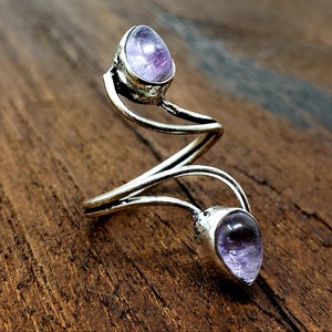 Può includere: Un anello argentato con due pietre di ametista viola a forma di goccia. L'anello ha un design aperto unico che si avvolge attorno al dito. Le pietre preziose sono incastonate in un'incastonatura a castone e l'anello è esposto su una superficie di legno.