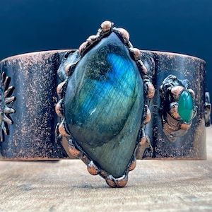 May include: A wide copper cuff bracelet featuring a large, iridescent labradorite stone at the center. The bracelet has a textured finish and is adorned with a smaller green stone and sunburst designs. A unique piece of jewelry.