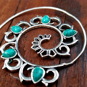 May include: Silver-toned spiral earrings with turquoise teardrop-shaped stones. The earrings have a detailed, openwork design with a central spiral element. The earrings are displayed on a wooden surface.