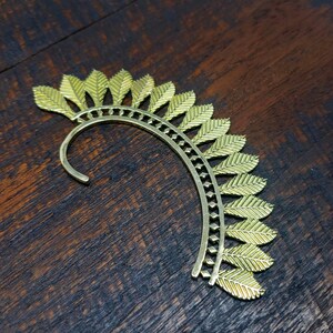 May include: Gold ear cuff with a leaf design. The cuff is curved and has a delicate, intricate pattern.
