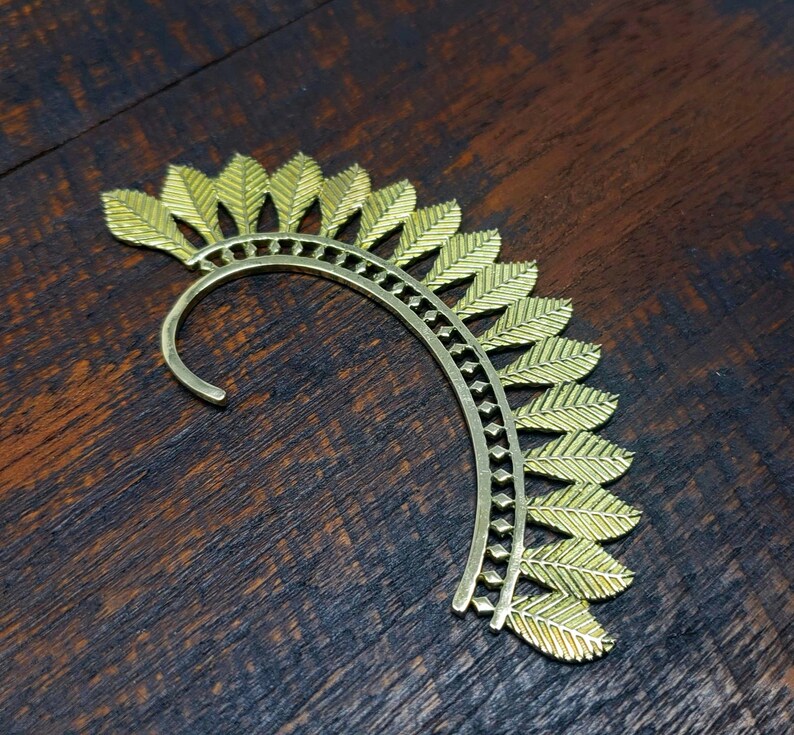 May include: Gold ear cuff with a leaf design. The cuff is curved and has a delicate, intricate pattern.