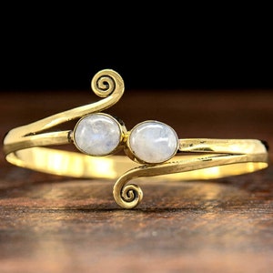 May include: A gold-toned bracelet featuring two white moonstone cabochons. The bracelet has a delicate, swirling design.