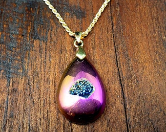 Peut inclure: Un collier avec un pendentif en forme de larme sur une chaîne dorée. Le pendentif est en agate violette irisée avec un amas de cristaux bleu foncé et or à l'intérieur.