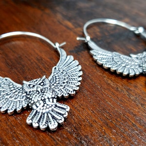 May include: A pair of silver hoop earrings with an owl design. The owls have detailed wings and are facing forward with their eyes open.
