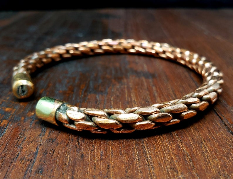 May include: A copper bracelet with a braided design. The bracelet is open and has a smooth, rounded edge.