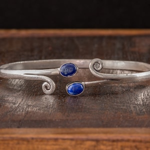 以下が含まれることがあります： 2つの青い楕円形の石が付いたシルバーのブレスレット。ブレスレットは、中央で交わる2本の曲線でシンプルなデザインになっています。