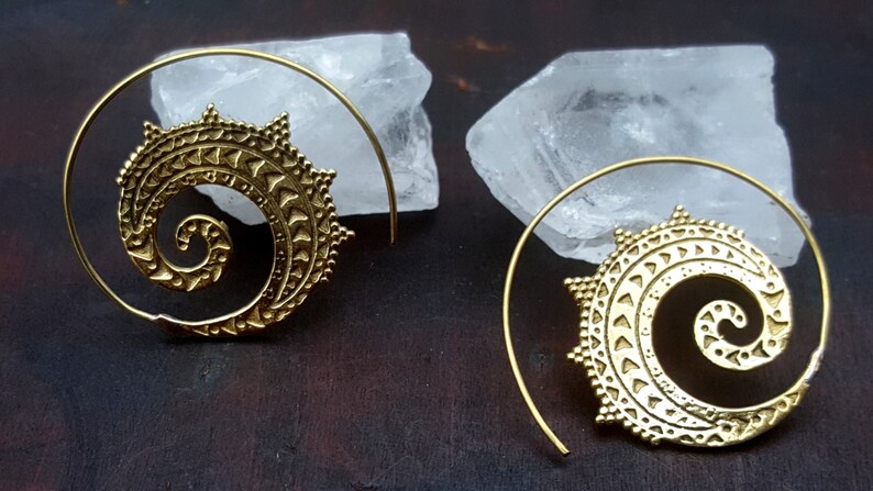 May include: A pair of gold-toned spiral hoop earrings with a detailed, intricate design. The earrings are made of metal and have a circular shape.