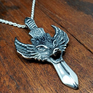 May include: A silver-colored pendant necklace featuring a wolf head design with a sword. The wolf's head is detailed with intricate carvings, and the sword blade extends downwards. The necklace chain is a silver rope style.