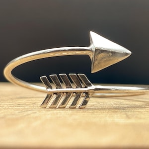 May include: A silver arrow bracelet. The arrow head is triangular, and the fletching has multiple lines. The bracelet curves around. The background is a wooden surface. The bracelet is a unique design.