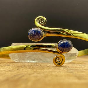 May include: A gold-colored cuff bracelet with a spiral design at each end. The bracelet features two round, dark blue gemstones. The jewelry is displayed on a clear, crystalline object, set against a dark background.