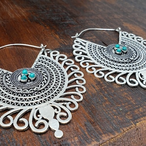 May include: Pair of silver-toned hoop earrings with intricate, openwork designs. Each earring features a semi-circular shape with a central circular element adorned with three small turquoise-colored stones. The earrings have a detailed, bohemian aesthetic.