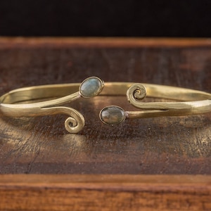 May include: A gold-colored cuff bracelet with two oval-shaped, gray-green gemstones. The bracelet has a curved design with decorative swirls at each end. The jewelry is displayed on a dark wooden surface.