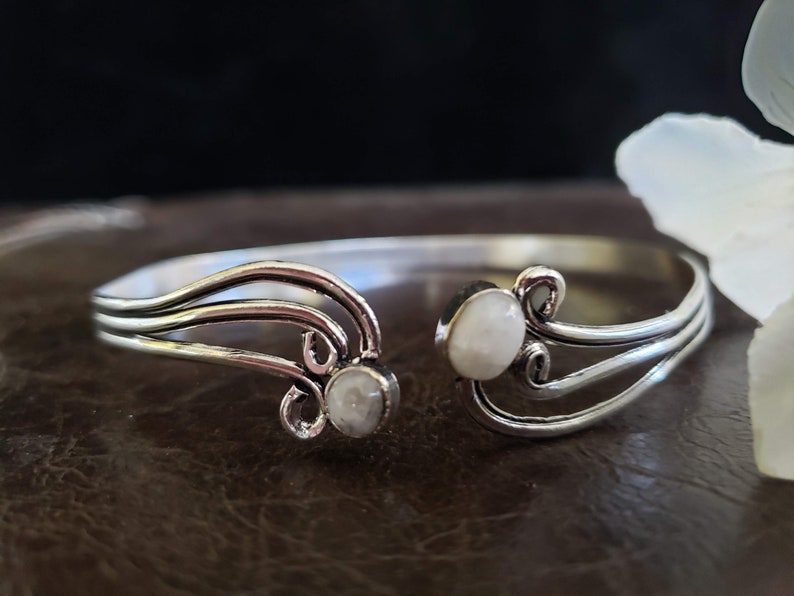 K&ouml;nnte beinhalten: Silbernes Armreif-Armband mit zwei wei&szlig;en Steinen, die in das Design eingefasst sind. Das Armband hat ein zartes, flie&szlig;endes Design.