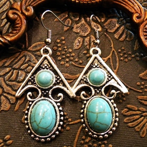 May include: A pair of silver-toned earrings with turquoise-colored oval and round stones. The earrings have a triangular top design with a smaller stone and an oval stone at the bottom, surrounded by a beaded border. The earrings are designed with a vintage or bohemian style.