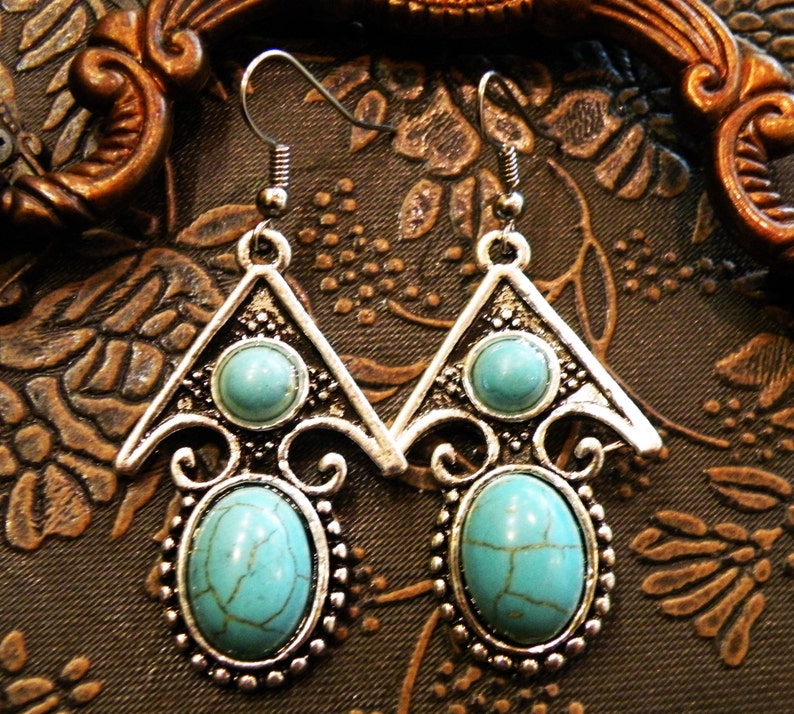 May include: A pair of silver-toned earrings with turquoise-colored oval and round stones. The earrings have a triangular top design with a smaller stone and an oval stone at the bottom, surrounded by a beaded border. The earrings are designed with a vintage or bohemian style.