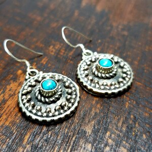 May include: A pair of silver-toned earrings with a turquoise stone center. The earrings have a circular design with a beaded border and a smaller turquoise stone in the center. The earrings are on a dark wooden surface.