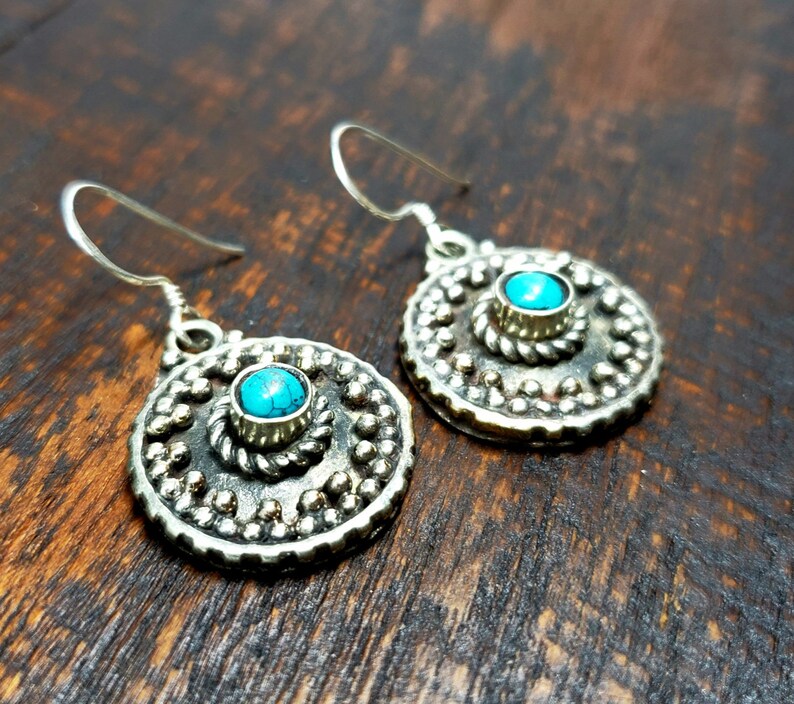 May include: A pair of silver-toned earrings with a turquoise stone center. The earrings have a circular design with a beaded border and a smaller turquoise stone in the center. The earrings are on a dark wooden surface.