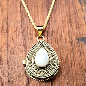 May include: A gold teardrop-shaped locket necklace with a white stone in the center. The locket has intricate details and a rope chain.