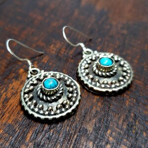 May include: A pair of silver-toned earrings with a turquoise stone center. The earrings have a circular design with a beaded border and a twisted rope detail around the stone. The earrings are displayed on a dark wooden surface.