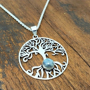 May include: A silver tree of life pendant necklace with a blue labradorite gemstone in the center. The pendant is round and features a detailed design of a tree with branches and roots.