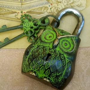 May include: A green and black metal owl padlock with a silver keyhole and two keys. The owl's eyes are green and have a swirling pattern.